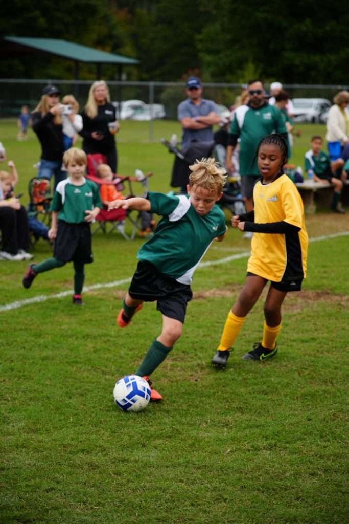 boy-kicking-ball-18613939 Boys and girls in team soccer jerseys playing soccer on a green field outdoors.